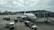 A large passenger airplane is parked at an airport gate, connected to a jet bridge. Several ground service vehicles and workers are visible, performing various operations around the aircraft. In the background, another airplane is visible on the runway, with a backdrop of distant hills and buildings under a cloudy sky.