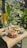 A woven mat on a small table holds several offerings consisting of fruits, colorful flowers, and handcrafted decorations. The setup is outdoors, surrounded by lush, tropical greenery with large leaves and a tree trunk wrapped in yellow fabric.