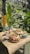 A woven mat on a small table holds several offerings consisting of fruits, colorful flowers, and handcrafted decorations. The setup is outdoors, surrounded by lush, tropical greenery with large leaves and a tree trunk wrapped in yellow fabric.