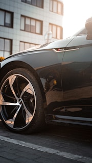 A close-up of a sleek black car's front wheel and side badge, with a building in the background. The wheel design is modern and sporty, and the car exhibits a polished finish.