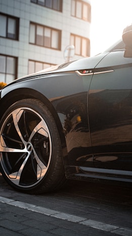 A close-up of a sleek black car's front wheel and side badge, with a building in the background. The wheel design is modern and sporty, and the car exhibits a polished finish.