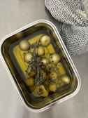 A rectangular tray containing pickled vegetables submerged in oil along with herbs and peppercorns. The tray is made of metal and sits on a smooth, gray surface. Beside the tray, there is a patterned cloth with a black and white weave.