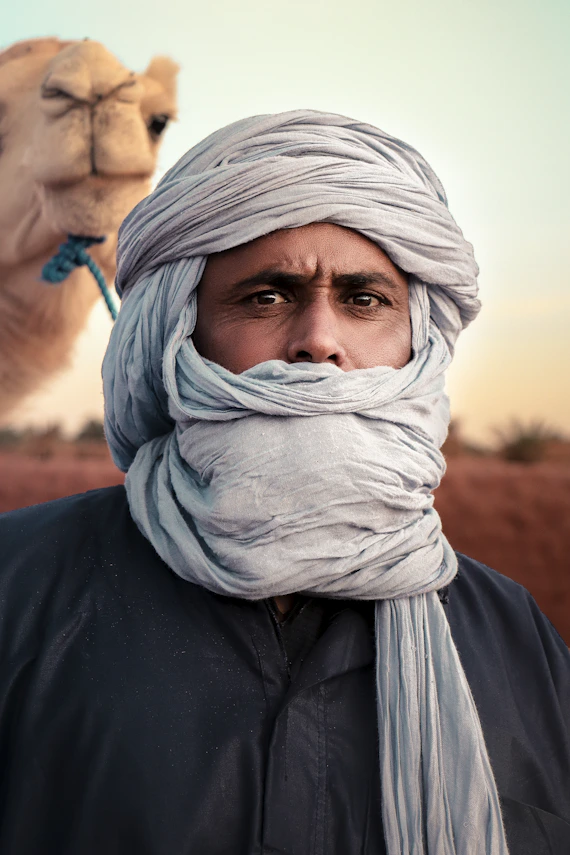 Guide touareg portant un turban traditionnel, avec un chameau en arrière-plan lors du trek vers Sefa