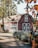 A rustic red barn with white trim is situated in a tranquil setting surrounded by trees and lush greenery. The barn has large windows and a gambrel roof, with a garden featuring various plants and flowers nearby. Sunlight filters through the leaves, casting gentle shadows on the ground.