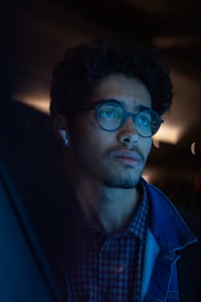 A man with curly hair wearing glasses and a plaid shirt, listening to music with wireless earbuds, gazes contemplatively into the distance. The lighting casts a cool blue tone over his face, creating a calm and introspective mood.