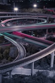 A complex network of elevated highways at night, featuring intertwining loops and illuminated by streetlights. The scene is vibrant with red and blue light trails from moving vehicles, reflecting the busy and dynamic nature of the city.