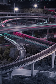 A complex network of elevated highways at night, featuring intertwining loops and illuminated by streetlights. The scene is vibrant with red and blue light trails from moving vehicles, reflecting the busy and dynamic nature of the city.