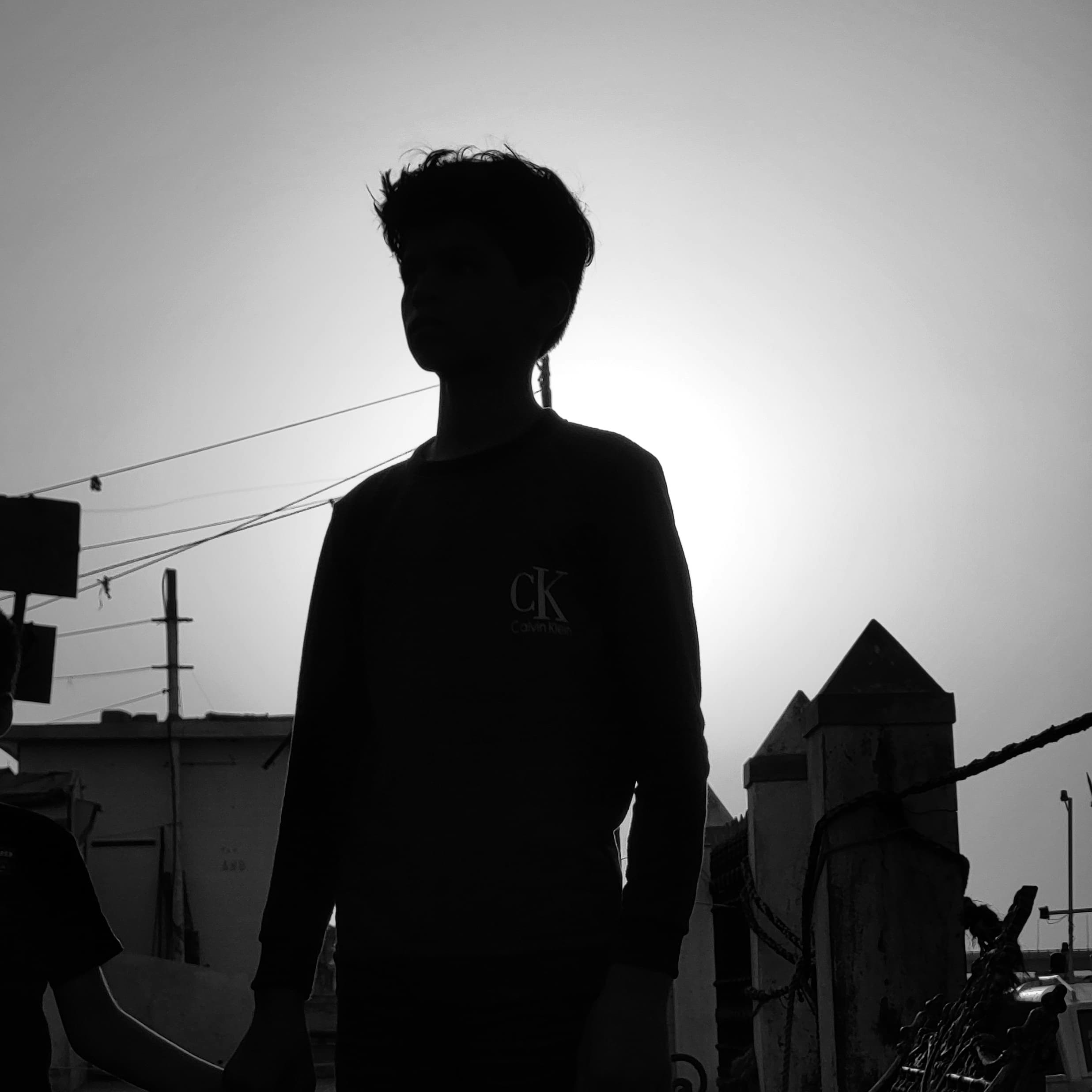 Black and white silhouette of young man, power lines and fence behind him. 