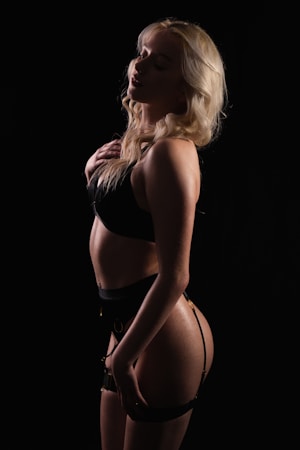 A person with long, blonde hair stands in profile against a black background, wearing dark lingerie. The lighting is low key, highlighting the contours and muscles of the person's body, creating a sense of depth and shadow.
