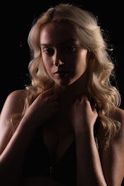 A person with long, wavy blonde hair appears in low key lighting, creating a dramatic contrast that emphasizes facial features and hair texture. The individual is holding their hands near their face, contributing to a pensive and moody ambiance.