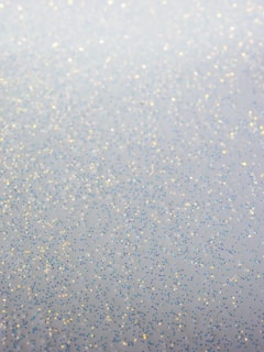 A soft and shimmering surface featuring scattered tiny glitter particles in various colors including blue, gold, and silver. The glitter appears to be sprinkled randomly across a blurred background, creating a subtle, delicate sparkle.