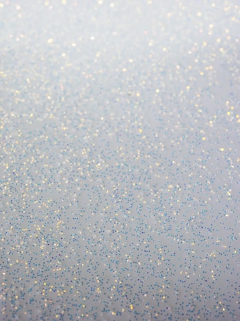 A soft and shimmering surface featuring scattered tiny glitter particles in various colors including blue, gold, and silver. The glitter appears to be sprinkled randomly across a blurred background, creating a subtle, delicate sparkle.