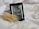 An e-reader displaying the cover of a book titled 'Sorcery of Thorns' by Margaret Rogerson rests on a soft, white fabric. A stalk of wheat is positioned beside the e-reader, adding a natural element to the scene. The color scheme of the e-book cover is primarily grayscale.