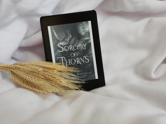 An e-reader displaying the cover of a book titled 'Sorcery of Thorns' by Margaret Rogerson rests on a soft, white fabric. A stalk of wheat is positioned beside the e-reader, adding a natural element to the scene. The color scheme of the e-book cover is primarily grayscale.