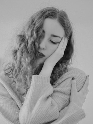 A black and white portrait of a young woman with curly hair. She rests her head on one hand, and her facial expression suggests deep thought or introspection. The other arm is wrapped around her body, creating a sense of self-comfort or contemplation. She wears a cozy sweater, and the overall composition is intimate and serene.