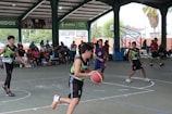A group of young basketball players are actively playing on an indoor court. The court is surrounded by a roofed structure with several spectators watching the game from the sidelines. The players are wearing team jerseys, and one of them is in the middle of dribbling the basketball. A mix of natural and artificial light illuminates the area, and a basketball hoop is visible in the background.