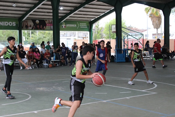 A group of young basketball players are actively playing on an indoor court. The court is surrounded by a roofed structure with several spectators watching the game from the sidelines. The players are wearing team jerseys, and one of them is in the middle of dribbling the basketball. A mix of natural and artificial light illuminates the area, and a basketball hoop is visible in the background.