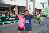 Two people in athletic clothing posing together at a public event, possibly a race or marathon. They are smiling and appear happy, with the person on the left holding up a peace sign. There are more people in the background, including children, and a banner displaying the word 'Apodaca'.