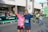 Two people in athletic clothing posing together at a public event, possibly a race or marathon. They are smiling and appear happy, with the person on the left holding up a peace sign. There are more people in the background, including children, and a banner displaying the word 'Apodaca'.