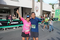 Two people in athletic clothing posing together at a public event, possibly a race or marathon. They are smiling and appear happy, with the person on the left holding up a peace sign. There are more people in the background, including children, and a banner displaying the word 'Apodaca'.