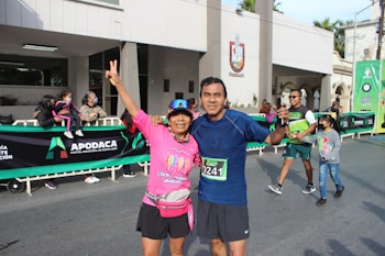 Two people in athletic clothing posing together at a public event, possibly a race or marathon. They are smiling and appear happy, with the person on the left holding up a peace sign. There are more people in the background, including children, and a banner displaying the word 'Apodaca'.