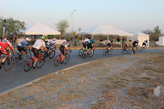 Cyclists are racing on a designated road, passing several white tents. The riders are in colorful jerseys and helmets, and they're going around a bend. A few spectators are visible behind a barricade.