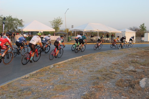 Cyclists are racing on a designated road, passing several white tents. The riders are in colorful jerseys and helmets, and they're going around a bend. A few spectators are visible behind a barricade.