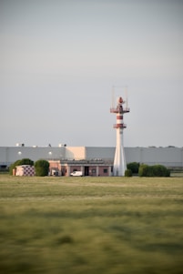 A large open grassy field with a white and red tower structure featuring an antenna on top. A smaller building with a white and red checkered pattern is situated nearby, along with several bushes. In the background, there is a large industrial building and a partially cloudy sky.