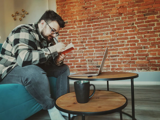A person wearing glasses and a checkered shirt is sitting on a teal sofa, focusing intently on writing in a notebook. In front of them, there is a small round wooden coffee table on which a laptop is open, along with a black mug. The background features a textured red brick wall, creating a cozy and creative atmosphere.
