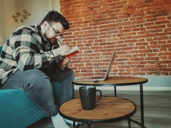 A person wearing glasses and a checkered shirt is sitting on a teal sofa, focusing intently on writing in a notebook. In front of them, there is a small round wooden coffee table on which a laptop is open, along with a black mug. The background features a textured red brick wall, creating a cozy and creative atmosphere.