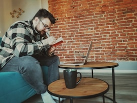 A person wearing glasses and a checkered shirt is sitting on a teal sofa, focusing intently on writing in a notebook. In front of them, there is a small round wooden coffee table on which a laptop is open, along with a black mug. The background features a textured red brick wall, creating a cozy and creative atmosphere.