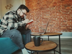 A person wearing glasses and a checkered shirt is sitting on a teal sofa, focusing intently on writing in a notebook. In front of them, there is a small round wooden coffee table on which a laptop is open, along with a black mug. The background features a textured red brick wall, creating a cozy and creative atmosphere.