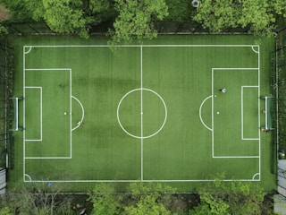 An aerial view of a green soccer field surrounded by trees, displaying marked white lines for gameplay. In each goal area, a player is positioned, while another player stands near the center circle. The field is framed by a fence, providing a clear boundary.