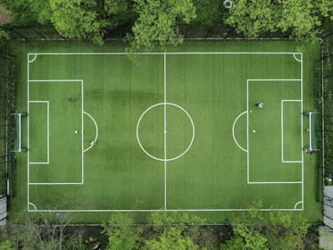 An aerial view of a green soccer field surrounded by trees, displaying marked white lines for gameplay. In each goal area, a player is positioned, while another player stands near the center circle. The field is framed by a fence, providing a clear boundary.
