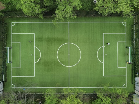 An aerial view of a green soccer field surrounded by trees, displaying marked white lines for gameplay. In each goal area, a player is positioned, while another player stands near the center circle. The field is framed by a fence, providing a clear boundary.