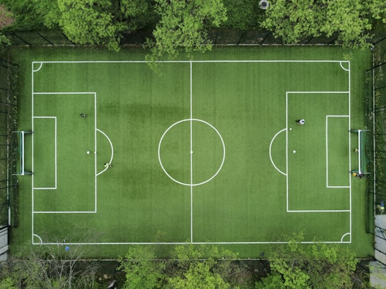 An aerial view of a green soccer field surrounded by trees, displaying marked white lines for gameplay. In each goal area, a player is positioned, while another player stands near the center circle. The field is framed by a fence, providing a clear boundary.