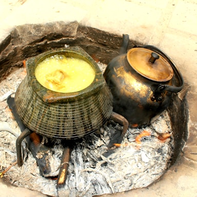 An outdoor cooking setup with two pots over a fire. One pot has a shiny, metallic surface and a lid, while the other is open and filled with a bubbling yellowish liquid, possibly soup or stew. The pots are positioned on a circular arrangement of charred wood and ashes.