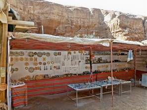 A small market stall set against a rocky backdrop displaying a variety of traditional and handmade items, including pottery, trinkets, and hanging decorations. The stall is adorned with colorful fabric and the items are neatly arranged on shelves and tables.