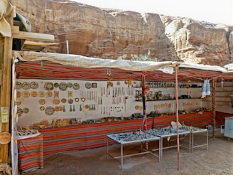 A small market stall set against a rocky backdrop displaying a variety of traditional and handmade items, including pottery, trinkets, and hanging decorations. The stall is adorned with colorful fabric and the items are neatly arranged on shelves and tables.