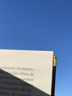 A book page is held up against a clear blue sky. The page contains text in Portuguese, with a shadow partially covering some of the words. The right edge of the book has a yellow fabric bookmark.