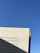 A book page is held up against a clear blue sky. The page contains text in Portuguese, with a shadow partially covering some of the words. The right edge of the book has a yellow fabric bookmark.
