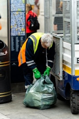 a man in a yellow safety vest and green gloves unloads a garbage bag
