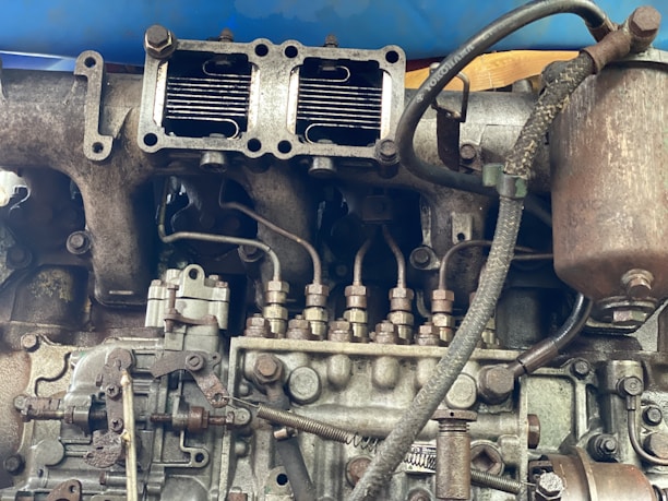 A detailed view of an engine part with various metallic components, including pipes, bolts, and connectors. The machinery appears old and worn, with visible rust and signs of usage. Several interconnected parts are assembled in a complex arrangement, suggesting industrial or automotive use.