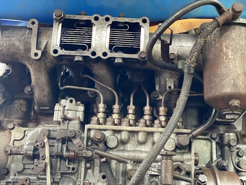 A detailed view of an engine part with various metallic components, including pipes, bolts, and connectors. The machinery appears old and worn, with visible rust and signs of usage. Several interconnected parts are assembled in a complex arrangement, suggesting industrial or automotive use.