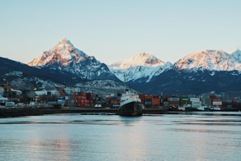 A serene harbor scene with a docked ship and stacked shipping containers, set against a backdrop of majestic snow-capped mountains in the distance. The water is calm, reflecting the hues of the sunset.