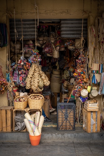A small shop is filled with a variety of handmade goods, including woven baskets, colorful textiles, and traditional crafts. The entrance is partially obscured by hanging ropes and items such as bags and hats. Wooden crates and a plastic basket are stacked on the ground, while bright tapestries and straw items add vibrancy to the display.