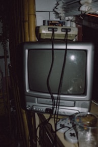 A retro television set with a gaming console on top, featuring multiple cables and controllers. The image includes a stack of papers and a glass placed beside the setup, with a dimly lit ambiance.