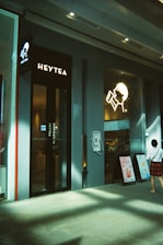 A modern tea shop featuring clean architectural lines and a minimalistic design. The brand name 'Heytea' is prominently displayed alongside a logo with a stylized person drinking from a cup. Posters advertising products are visible outside the entrance, and a person dressed casually walks by.