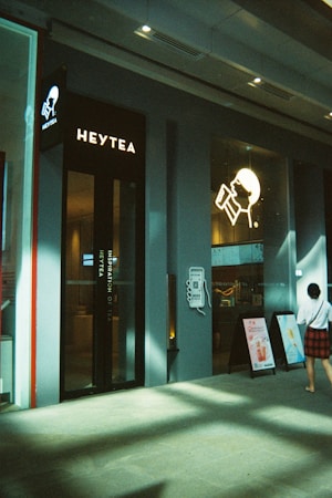 A modern tea shop featuring clean architectural lines and a minimalistic design. The brand name 'Heytea' is prominently displayed alongside a logo with a stylized person drinking from a cup. Posters advertising products are visible outside the entrance, and a person dressed casually walks by.