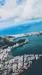 Aerial view of a coastal city with a large lagoon bordered by urban development. High-rise buildings and residential areas are nestled between lush green mountains and the ocean. The sky is partly cloudy, with patches of blue peeking through.